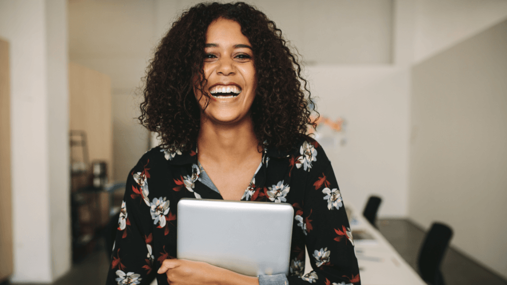 Young black female professional with curly hair standing in a conference room, holding a laptop and smiling warmly, embodying engaged leadership.