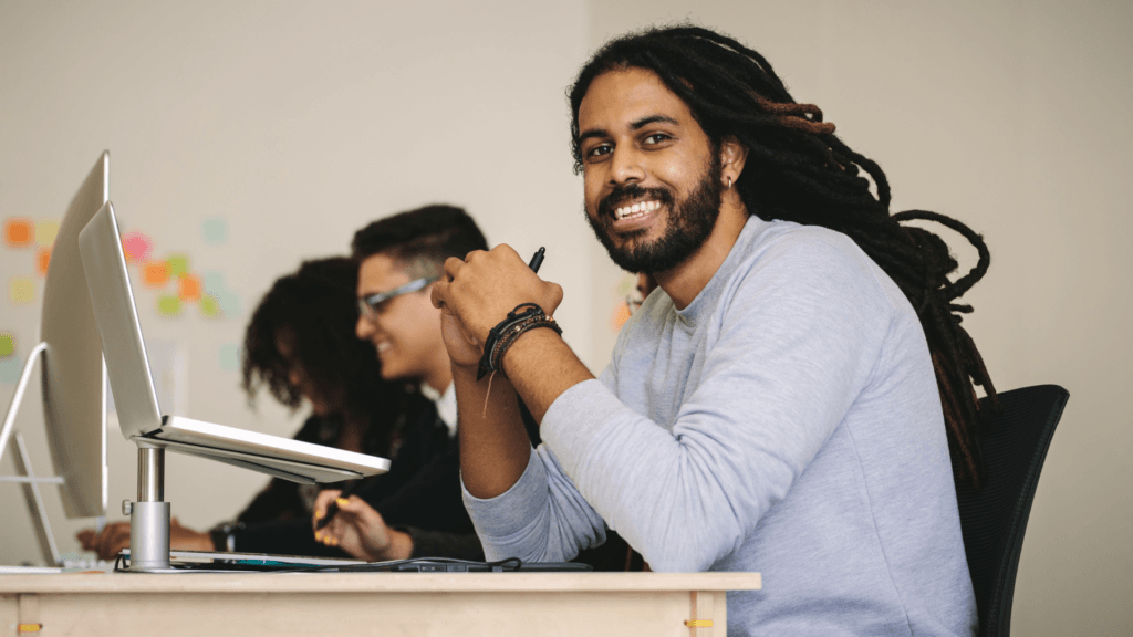 Smiling young professional with dreadlocks working at a modern office desk, using a digital tablet. A colleague is brainstorming in the background.